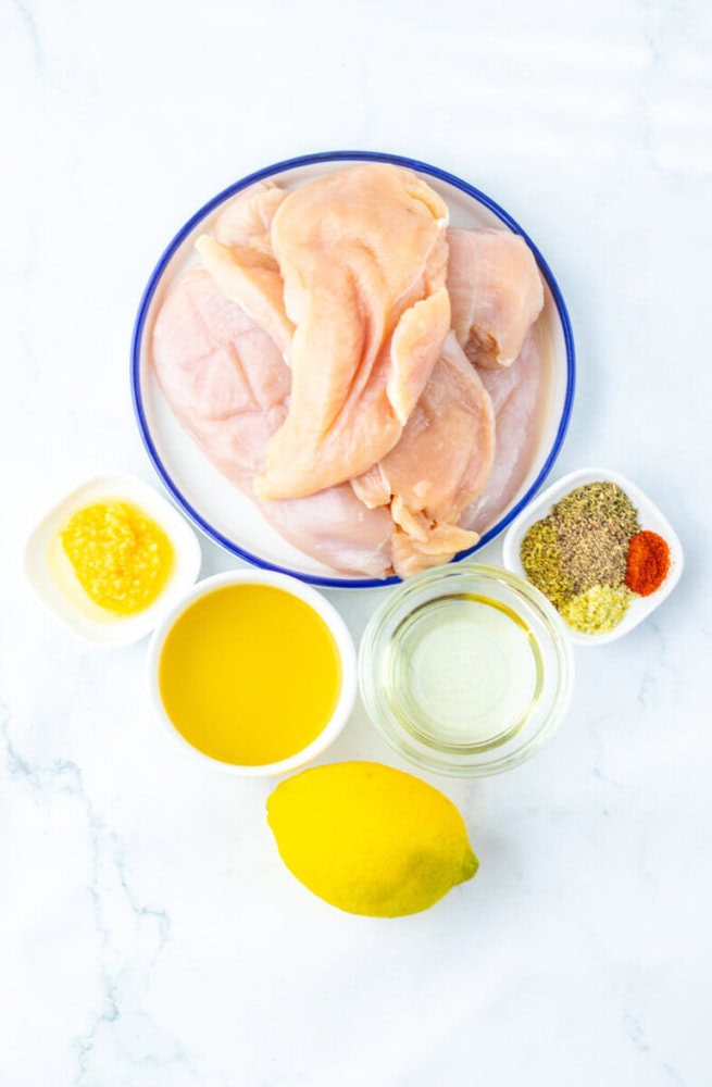 Raw chicken breasts on a plate, surrounded by small bowls of oil, melted butter, seasonings, herbs, and a whole lemon on a white surface.