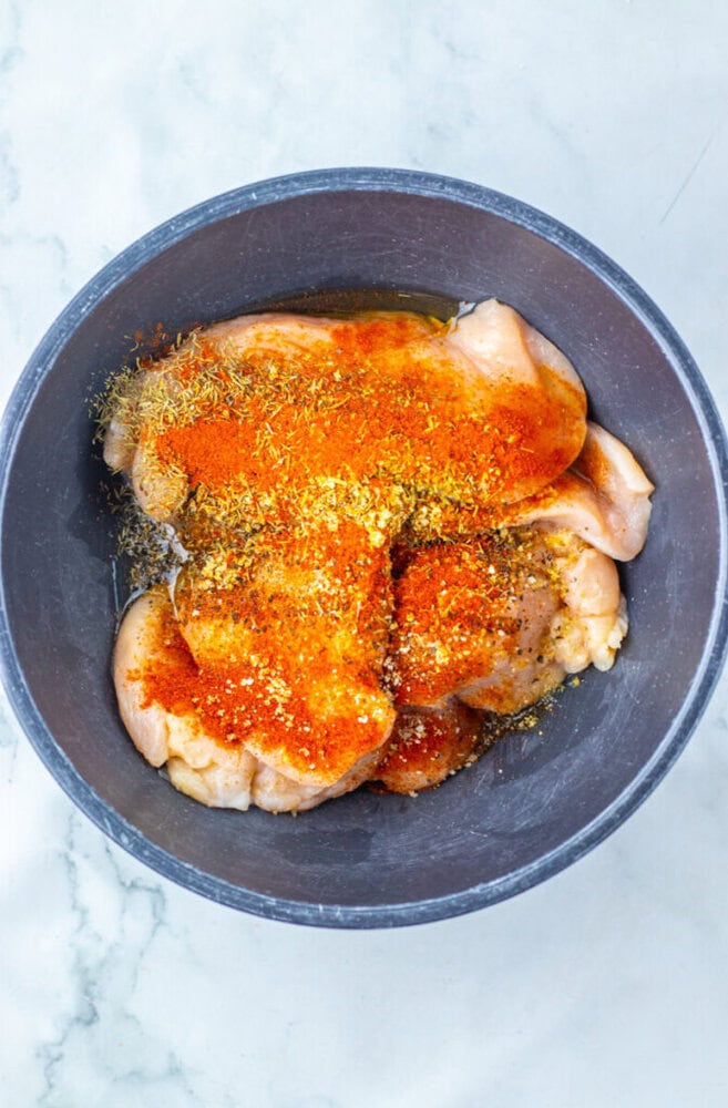 Raw chicken breasts in a black bowl, topped with assorted dry spices and seasoning, on a white marble surface.