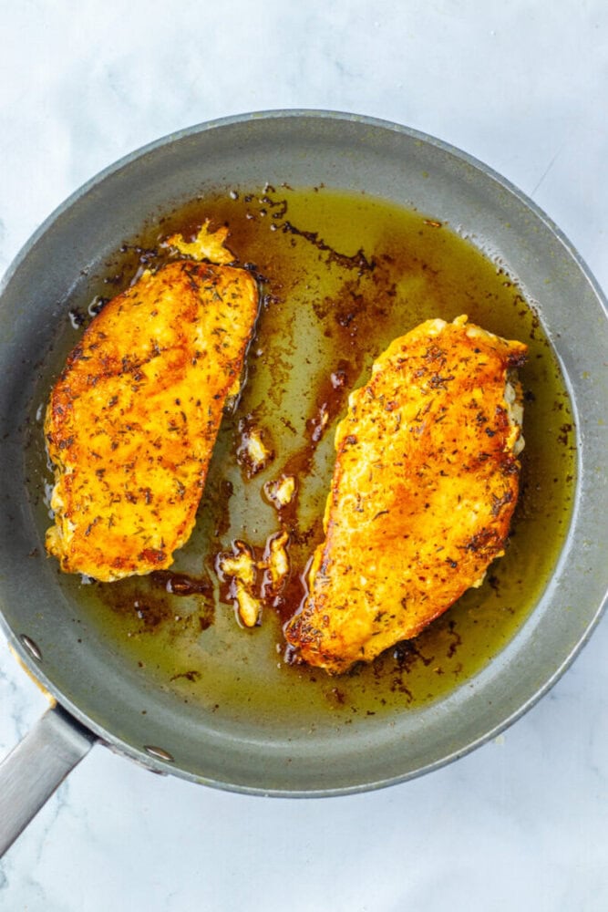 Two seasoned chicken breasts cooking in a frying pan with oil on a light-colored surface.