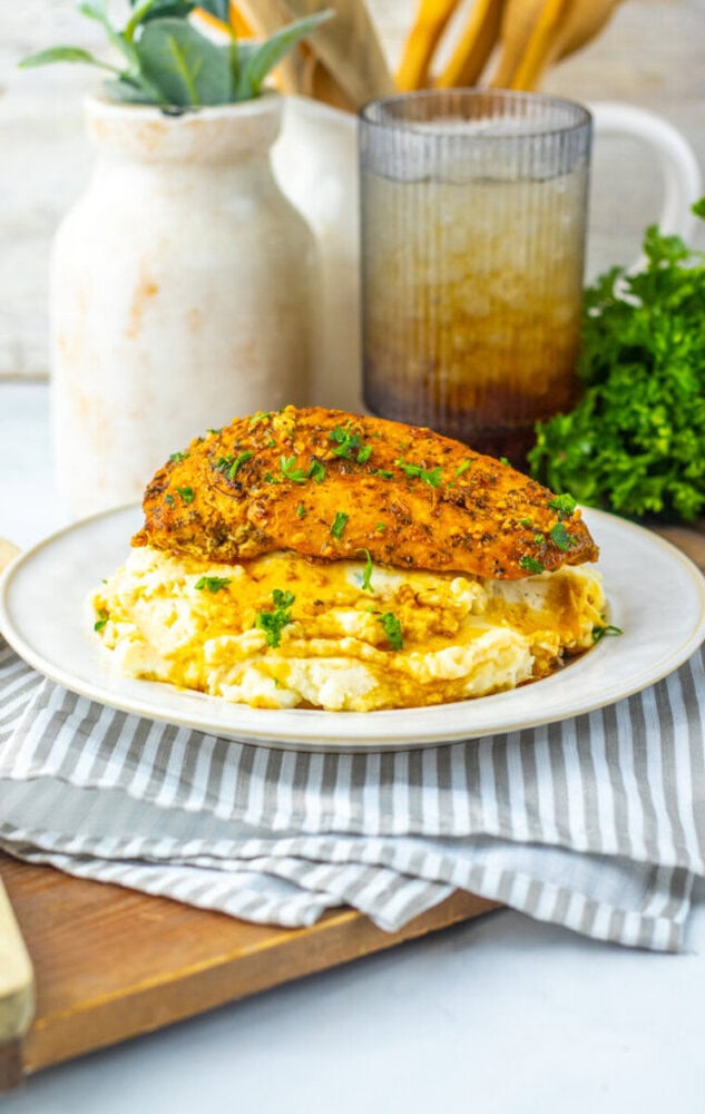 A plate of mashed potatoes topped with a seasoned, cooked chicken breast, garnished with chopped herbs, sits on a striped napkin.