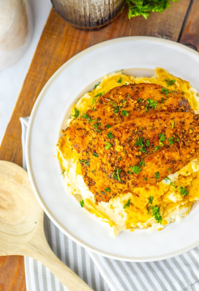 A seasoned, cooked chicken breast served on a bed of mashed potatoes, garnished with chopped parsley on a white plate.