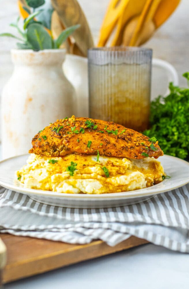 A seasoned, cooked chicken breast sits atop creamy mashed potatoes on a white plate, garnished with chopped herbs. Kitchen utensils and greenery are in the background.