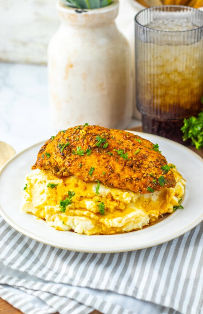 A seasoned chicken breast garnished with chopped herbs sits atop a serving of mashed potatoes on a white plate.
