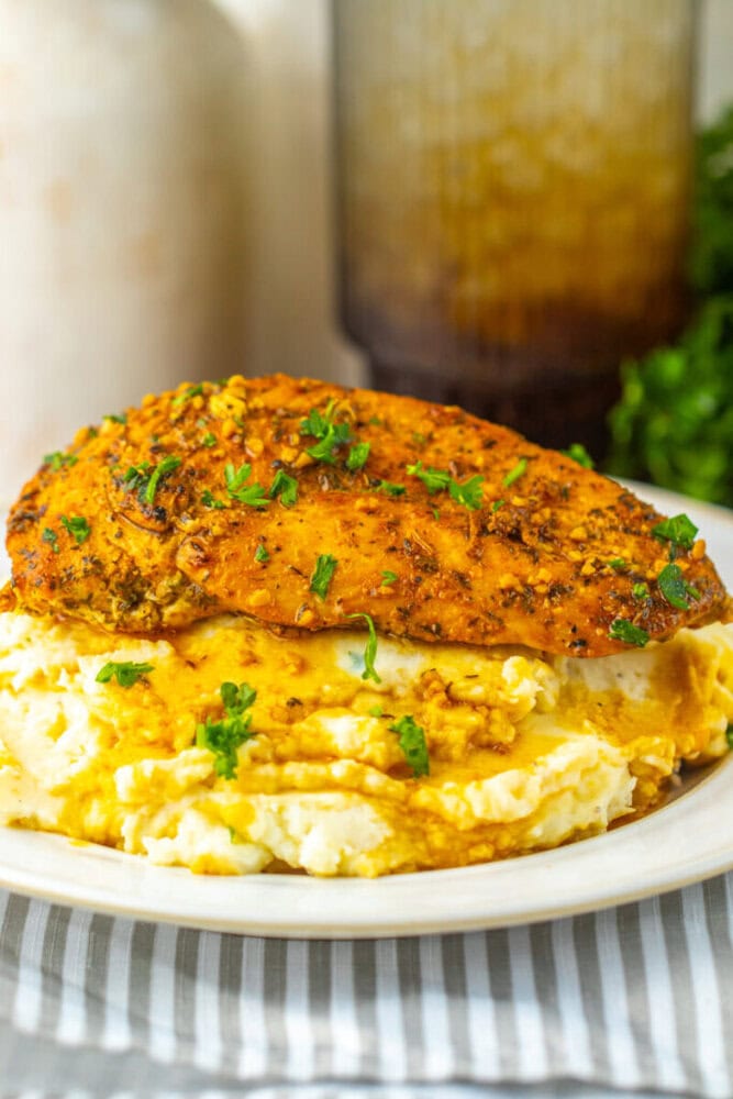 A seasoned baked chicken breast served on top of mashed potatoes with gravy, garnished with chopped parsley on a white plate.