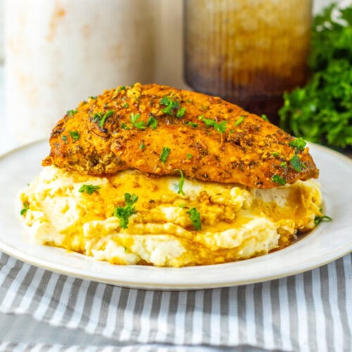 A plate with seasoned grilled chicken breast on top of mashed potatoes, garnished with chopped herbs, set on a striped napkin with a wooden spoon nearby.