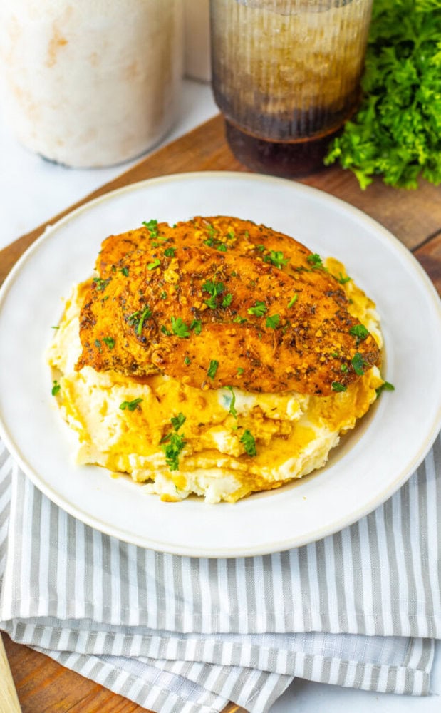 A seasoned, baked chicken breast served on a bed of mashed potatoes, garnished with chopped parsley on a white plate.