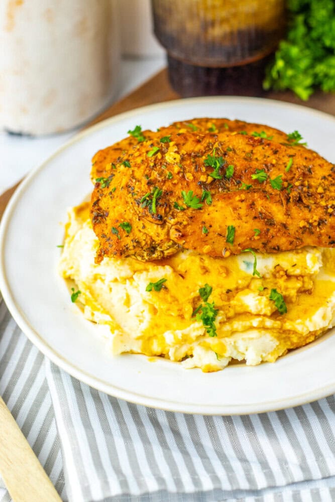 A seasoned, cooked chicken breast topped with chopped herbs sits on a bed of mashed potatoes with gravy on a white plate.