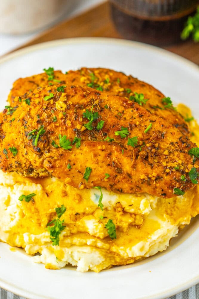 A seasoned baked chicken breast served on a bed of mashed potatoes, garnished with chopped parsley on a white plate.