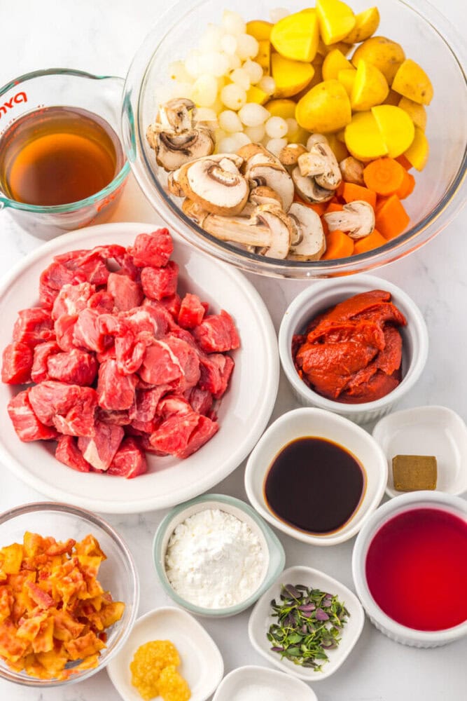 Various ingredients for beef stew are arranged in bowls, including cubed beef, mushrooms, pearl onions, potatoes, carrots, bacon, tomato paste, broth, soy sauce, wine, garlic, and seasonings.