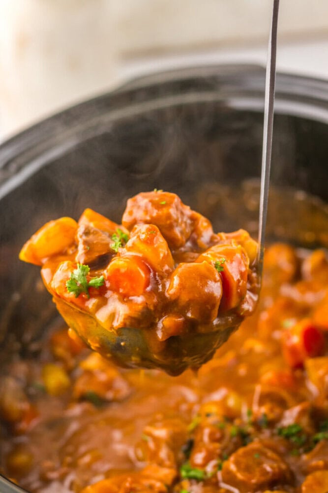 A ladle lifts a serving of beef stew with vegetables and thick brown sauce from a slow cooker.