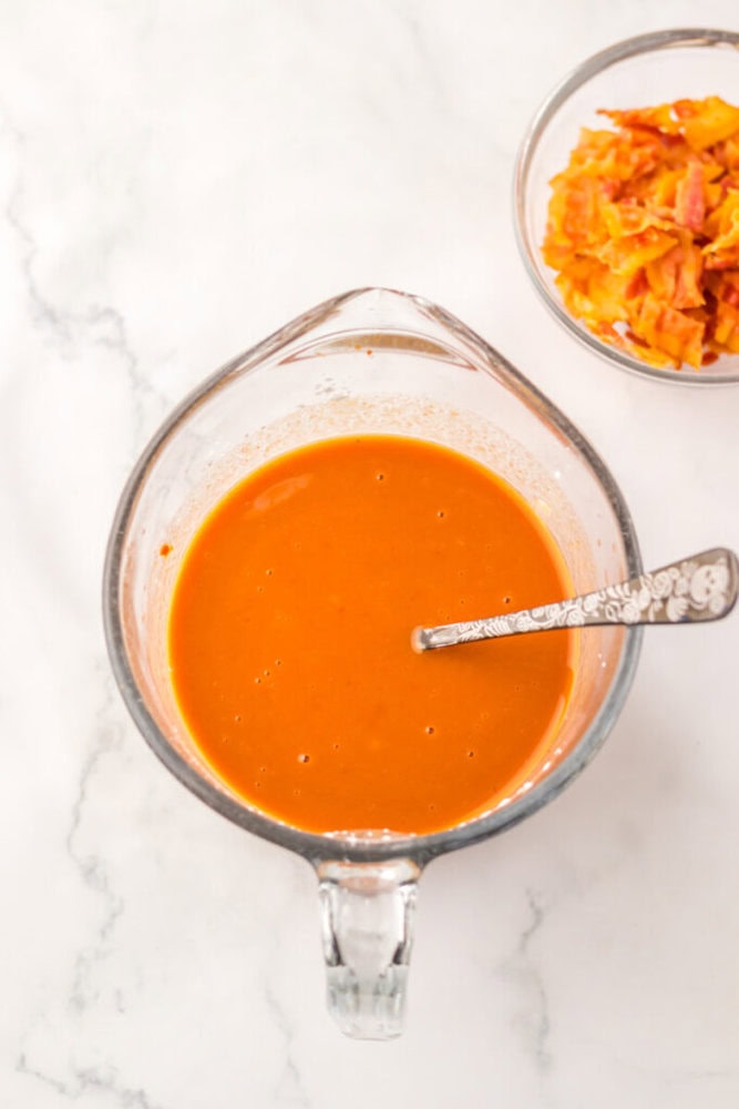 A glass measuring cup filled with orange sauce and a spoon, next to a small bowl containing orange vegetable peels, on a white marble surface.