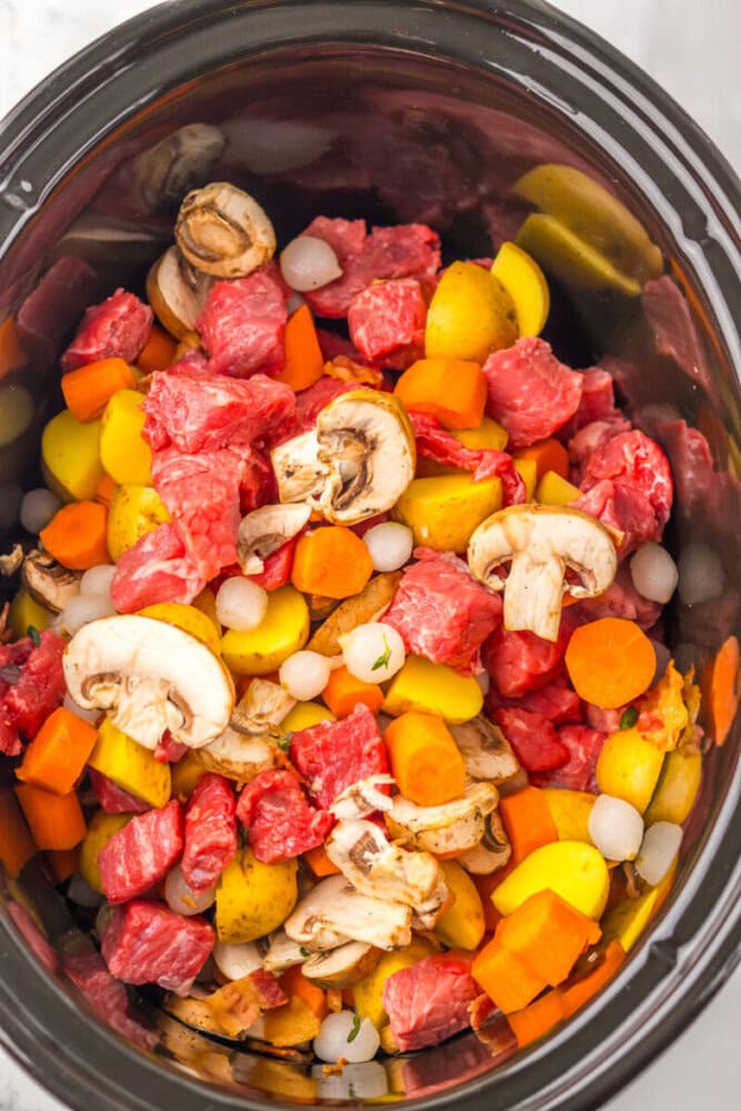 Chunks of raw beef, sliced mushrooms, carrots, potatoes, and pearl onions in a slow cooker, ready to be cooked.