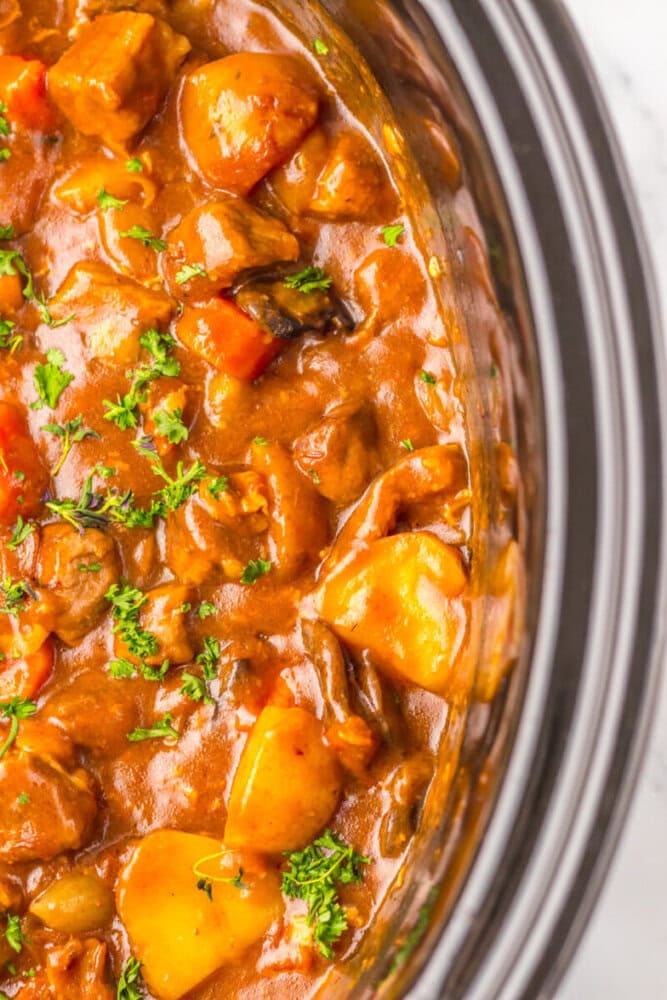 Close-up of beef stew with chunks of meat, potatoes, carrots, and parsley garnish in a slow cooker.