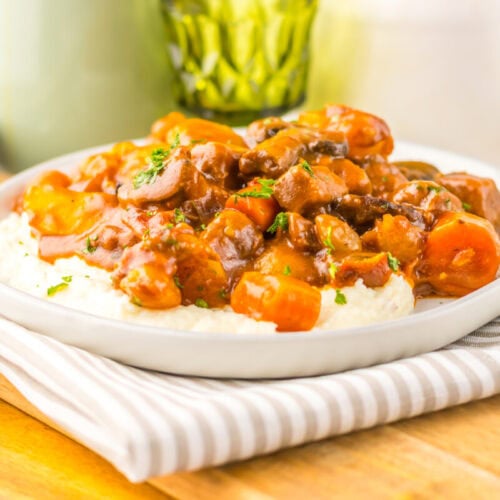 A plate of beef stew with carrots and sauce served over mashed potatoes, placed on a striped napkin with a wooden spoon nearby.