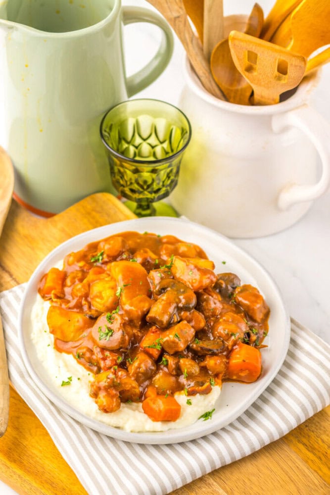 A plate of stew with carrots and sauce served over mashed potatoes, placed on a striped napkin next to a green glass, pitcher, and kitchen utensils.