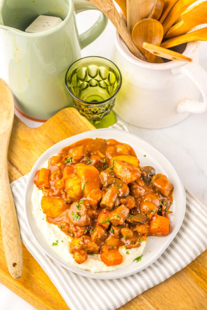 A plate of beef stew with potatoes and carrots served over mashed potatoes, placed on a striped napkin next to a wooden spoon and a green glass.