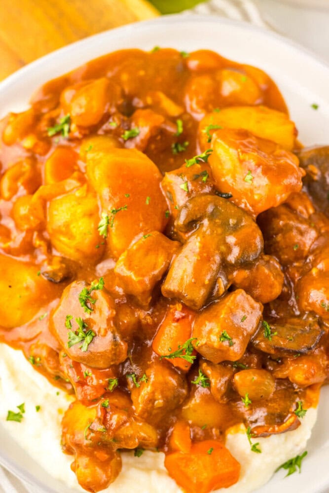 A plate of beef stew with carrots, potatoes, and mushrooms served over mashed potatoes, garnished with chopped parsley.
