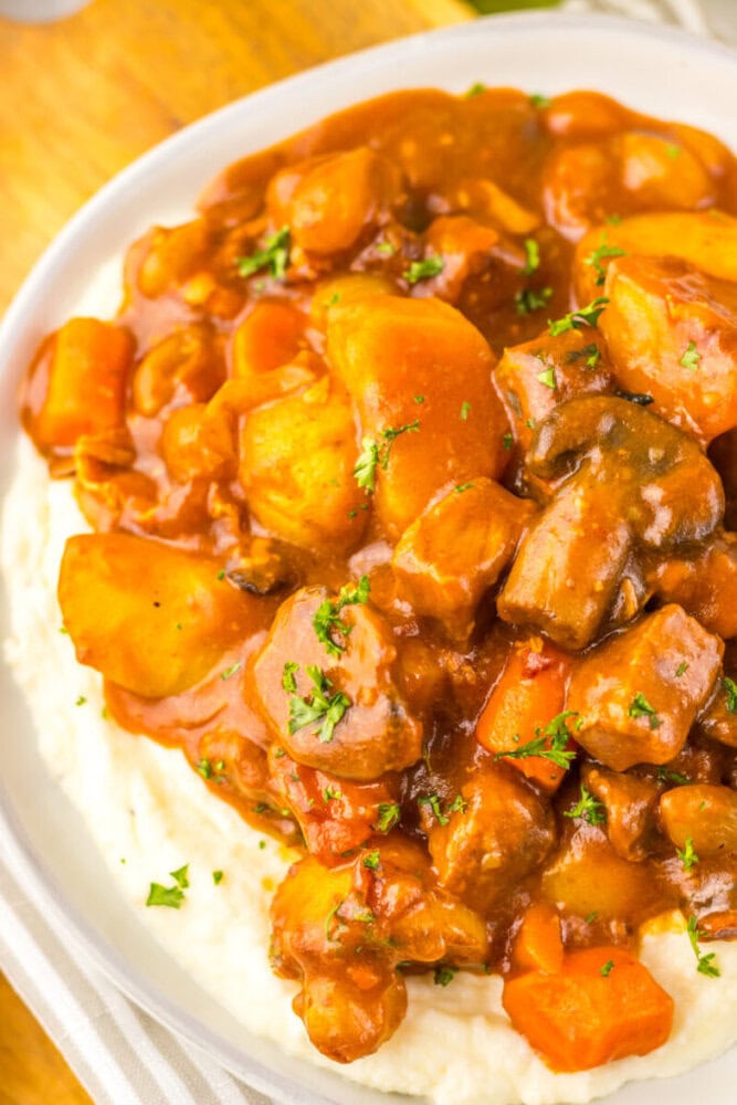 A plate of mashed potatoes topped with beef stew containing chunks of beef, potatoes, carrots, mushrooms, and garnished with chopped parsley.