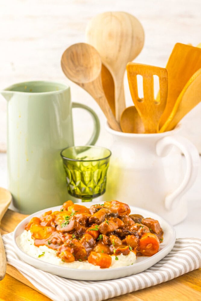 A plate of beef stew with carrots and sauce served over mashed potatoes, placed in front of a pitcher, glass, and a ceramic holder with wooden kitchen utensils.