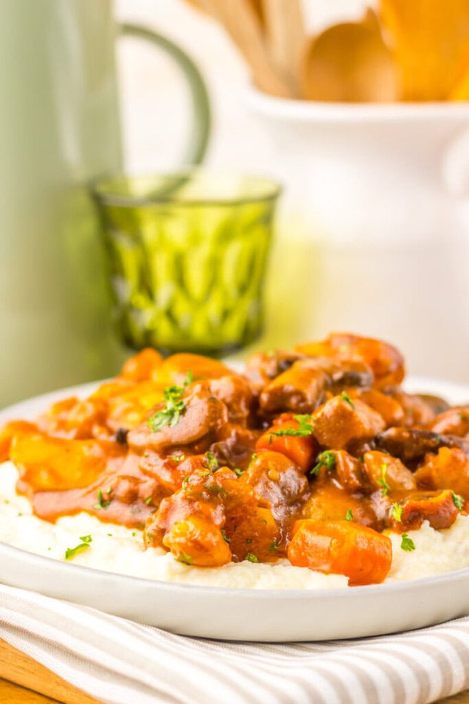 A plate of creamy mashed potatoes topped with a beef stew containing carrots and garnished with chopped herbs, with a green glass and kitchen utensils in the background.