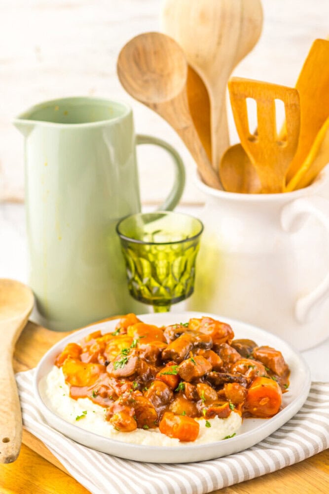 A plate of beef stew with carrots served over mashed potatoes sits on a striped napkin, with kitchen utensils, a green glass, and a pitcher in the background.