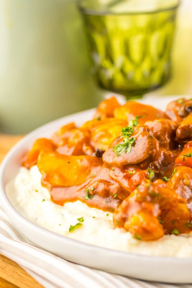 A plate of mashed potatoes topped with a beef and vegetable stew, garnished with chopped parsley. A green glass is in the background.