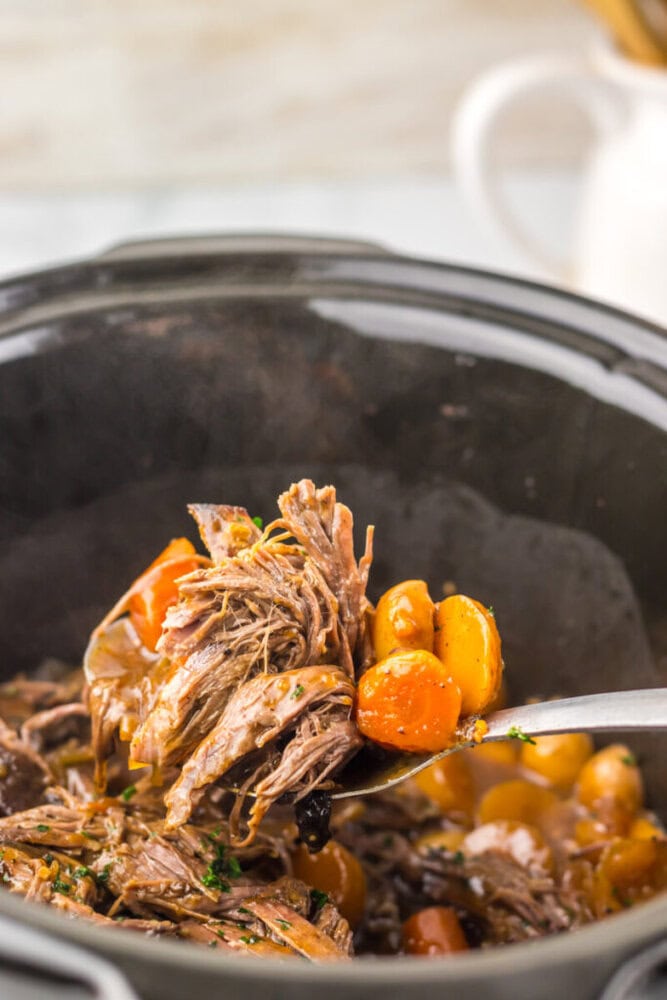 A spoon lifts shredded pot roast and cooked vegetables, including small potatoes and carrots, from a slow cooker.