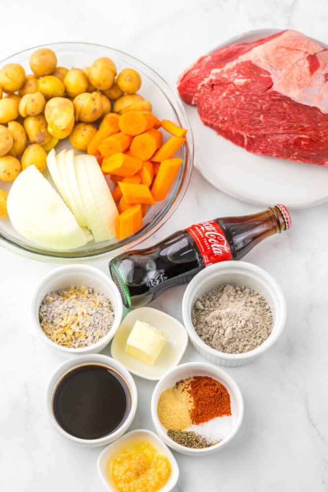 Ingredients for a beef pot roast are displayed, including raw beef, baby potatoes, carrots, onion, garlic, butter, spices, soup mix, soy sauce, flour, and a bottle of Coca-Cola.