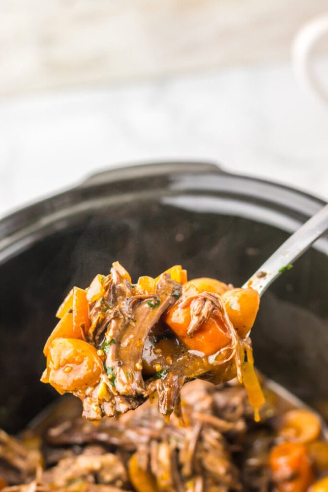 A close-up of a spoon lifting shredded beef, carrots, and vegetables in sauce from a slow cooker.