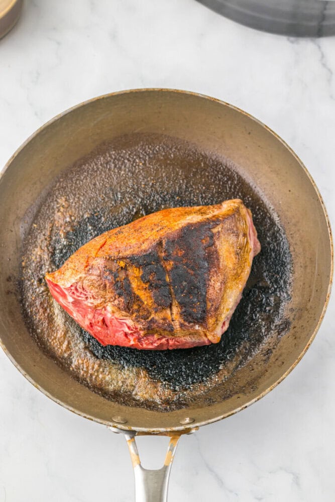 A piece of seasoned meat with a seared crust cooks in a skillet on a marble countertop.