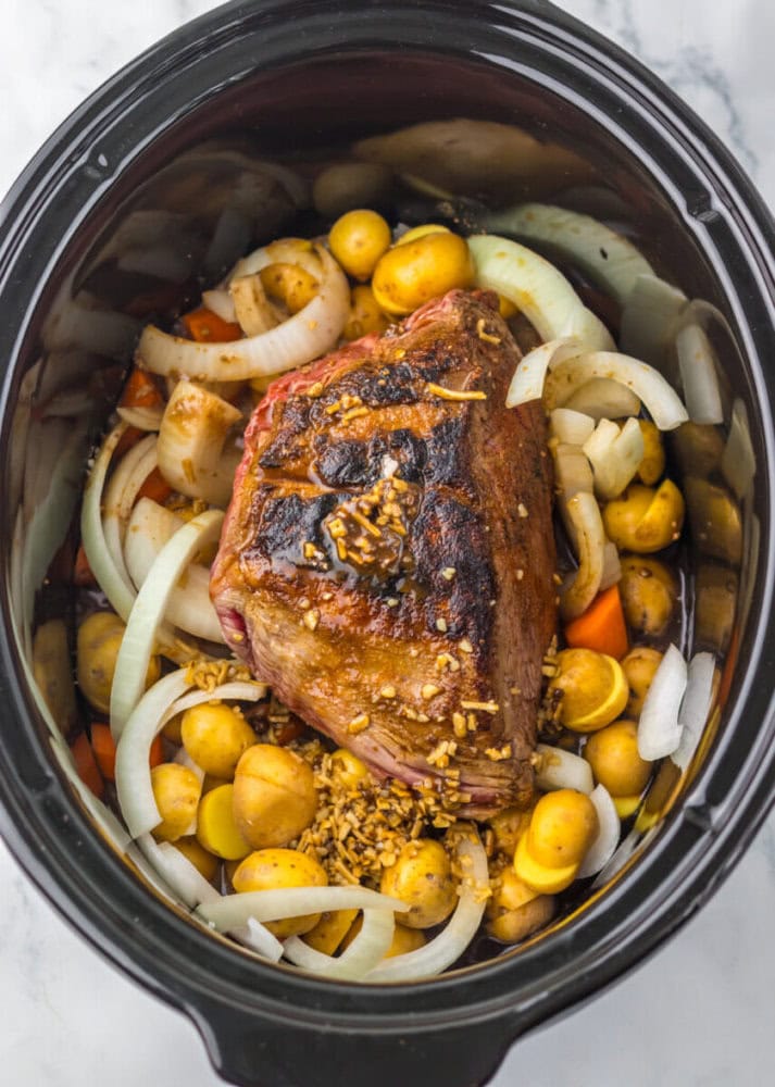 A browned roast with chopped onions, potatoes, carrots, and minced garlic sits in a slow cooker, ready to be cooked.