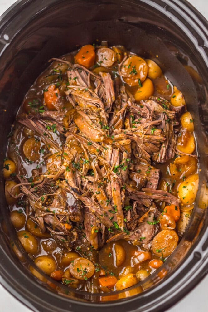 Shredded pot roast with carrots and baby potatoes in a brown gravy, served in a slow cooker.