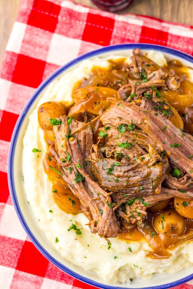 A plate of shredded pot roast with carrots and potatoes served over mashed potatoes, garnished with chopped herbs, on a red and white checkered tablecloth.