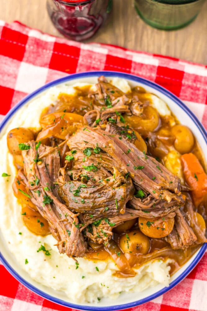 Shredded pot roast with carrots and potatoes served over creamy mashed potatoes on a white plate, placed on a red and white checkered tablecloth.