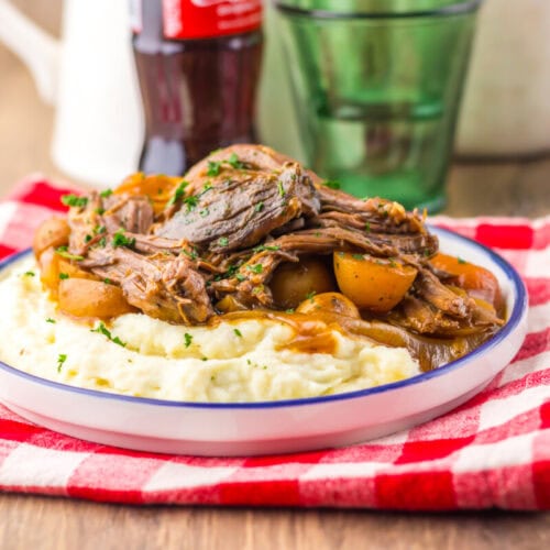 A plate of mashed potatoes topped with shredded pot roast, potatoes, and gravy sits on a red checkered cloth, with a glass and a soda bottle in the background.