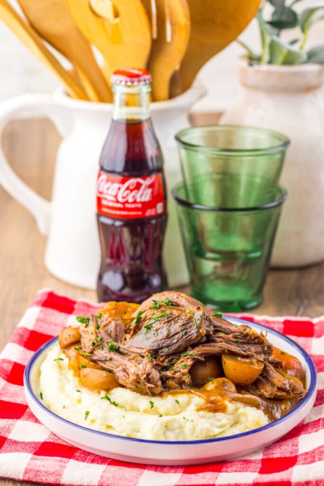 A plate with mashed potatoes, pot roast, and vegetables sits on a red checkered cloth beside a glass bottle of Coca-Cola, two green glasses, and kitchen utensils in the background.