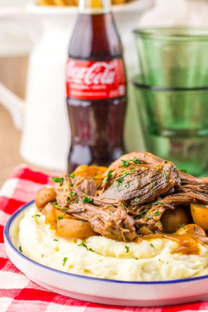 A plate of mashed potatoes topped with shredded pot roast and potatoes, garnished with herbs, sits in front of a glass bottle of Coca-Cola and green stacked glasses.