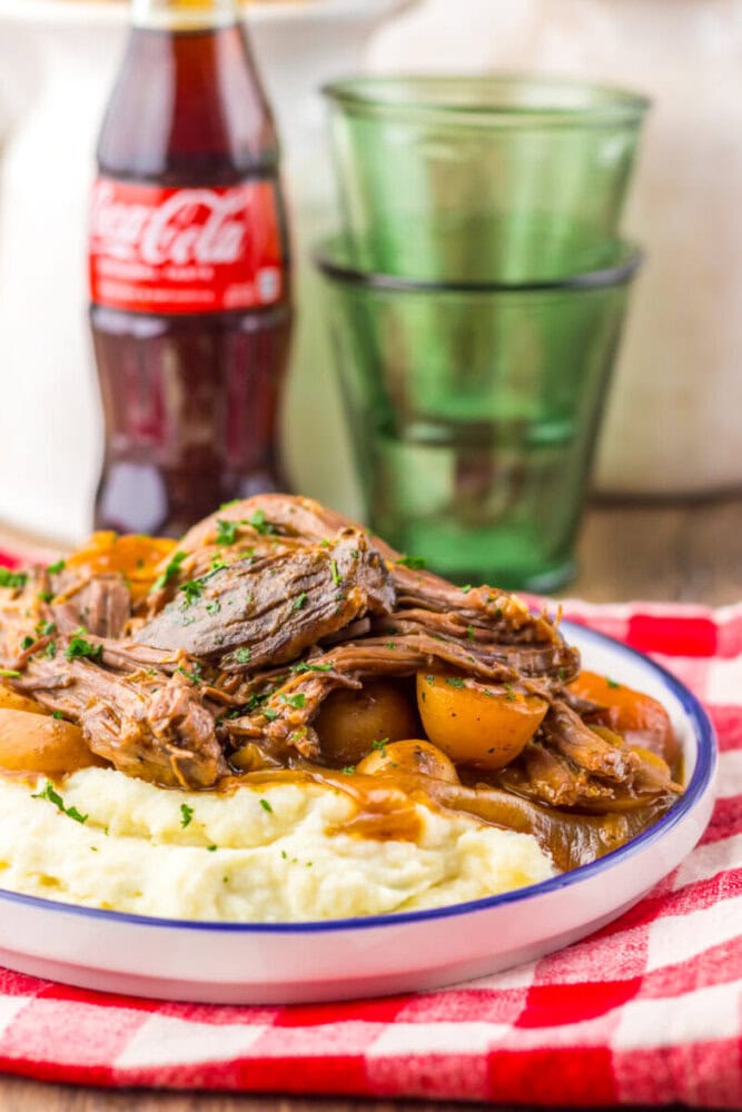 A plate of pot roast with potatoes and gravy served over mashed potatoes, with a bottle of Coca-Cola and stacked green glasses in the background.