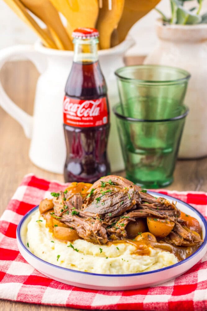 A plate of mashed potatoes topped with shredded pot roast and potatoes, placed on a red checkered cloth, with a glass Coke bottle and drinking glasses in the background.
