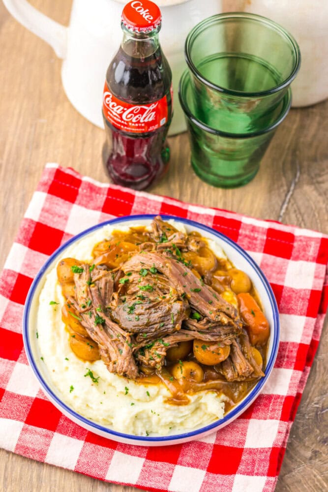 A plate of roast beef with mashed potatoes and vegetables on a red and white checkered cloth, next to a glass bottle of Coca-Cola and two empty green glasses.