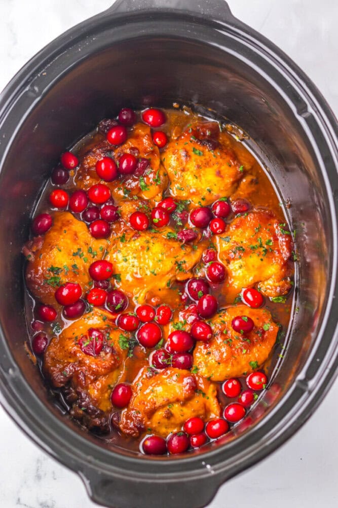 Chicken thighs cooked in a slow cooker with a sauce, garnished with whole cranberries and herbs.