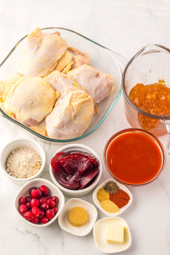 Raw chicken thighs in a glass dish, a measuring cup with orange sauce, and bowls with cranberries, cranberry sauce, spices, butter, and breadcrumbs on a white surface.
