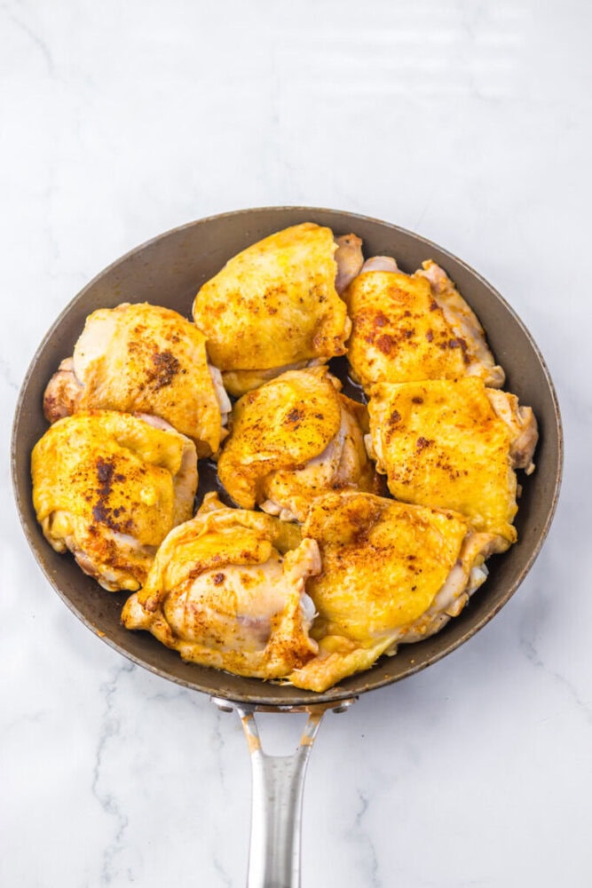 Seven seasoned, browned chicken thighs cooking in a skillet on a white marble surface.
