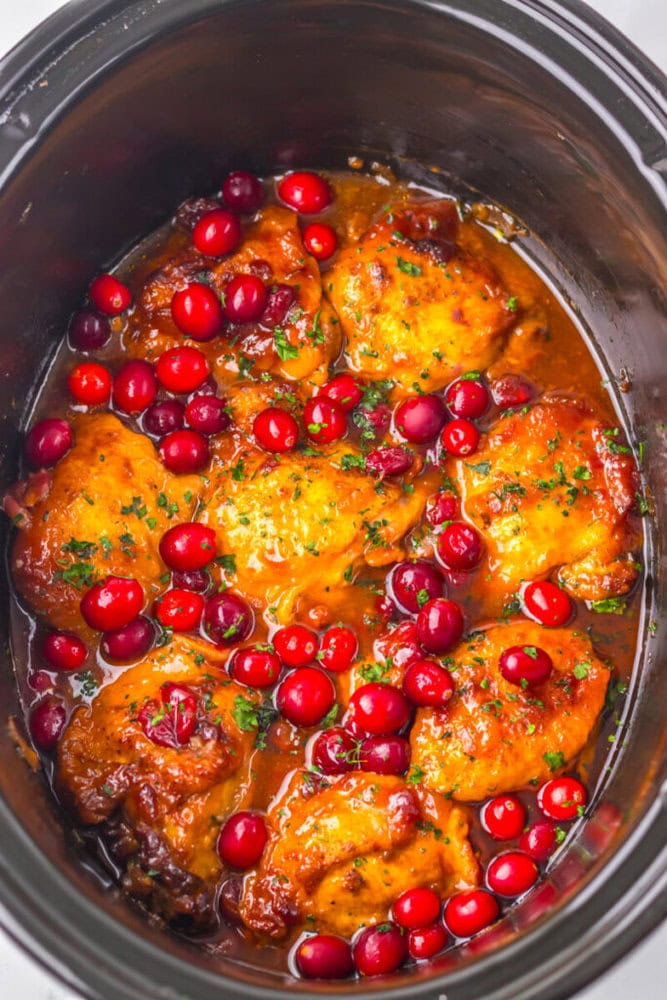 Chicken thighs cooked in a slow cooker with whole cranberries, coated in a glossy sauce and garnished with chopped herbs.