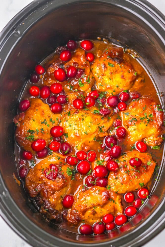 Chicken thighs cooked in a slow cooker with a sauce, garnished with whole cranberries and chopped herbs.