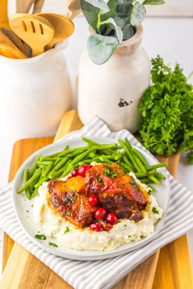 A plate of mashed potatoes topped with braised meat and cranberries, served with green beans, sits on a striped napkin next to a vase and a bunch of fresh parsley.