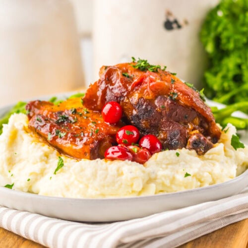 Braised meat served on mashed potatoes, garnished with cranberries and chopped herbs, with green beans and parsley in the background.