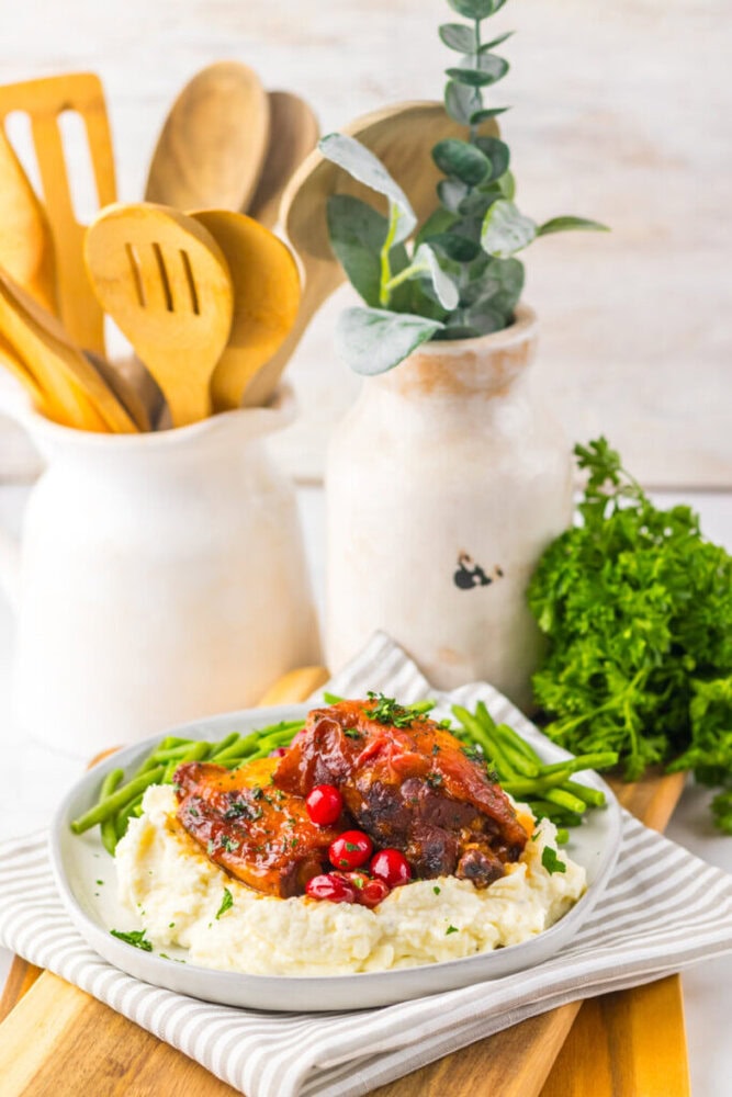 A plate of mashed potatoes topped with glazed meat and pomegranate seeds, served with green beans, with kitchen utensils and a vase in the background.