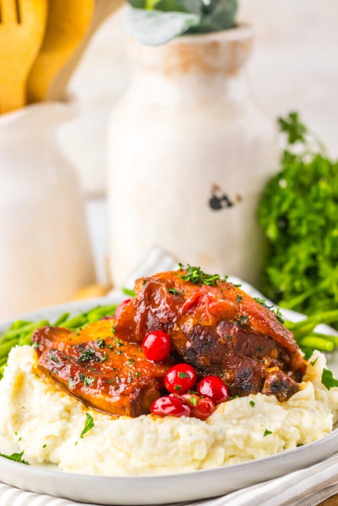 Braised meat with sauce and red berries served over mashed potatoes, garnished with chopped herbs, with green vegetables in the background.