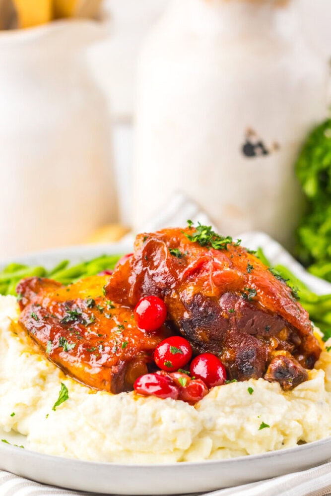 Braised meat served over mashed potatoes, garnished with fresh parsley and cranberries, with green beans in the background.